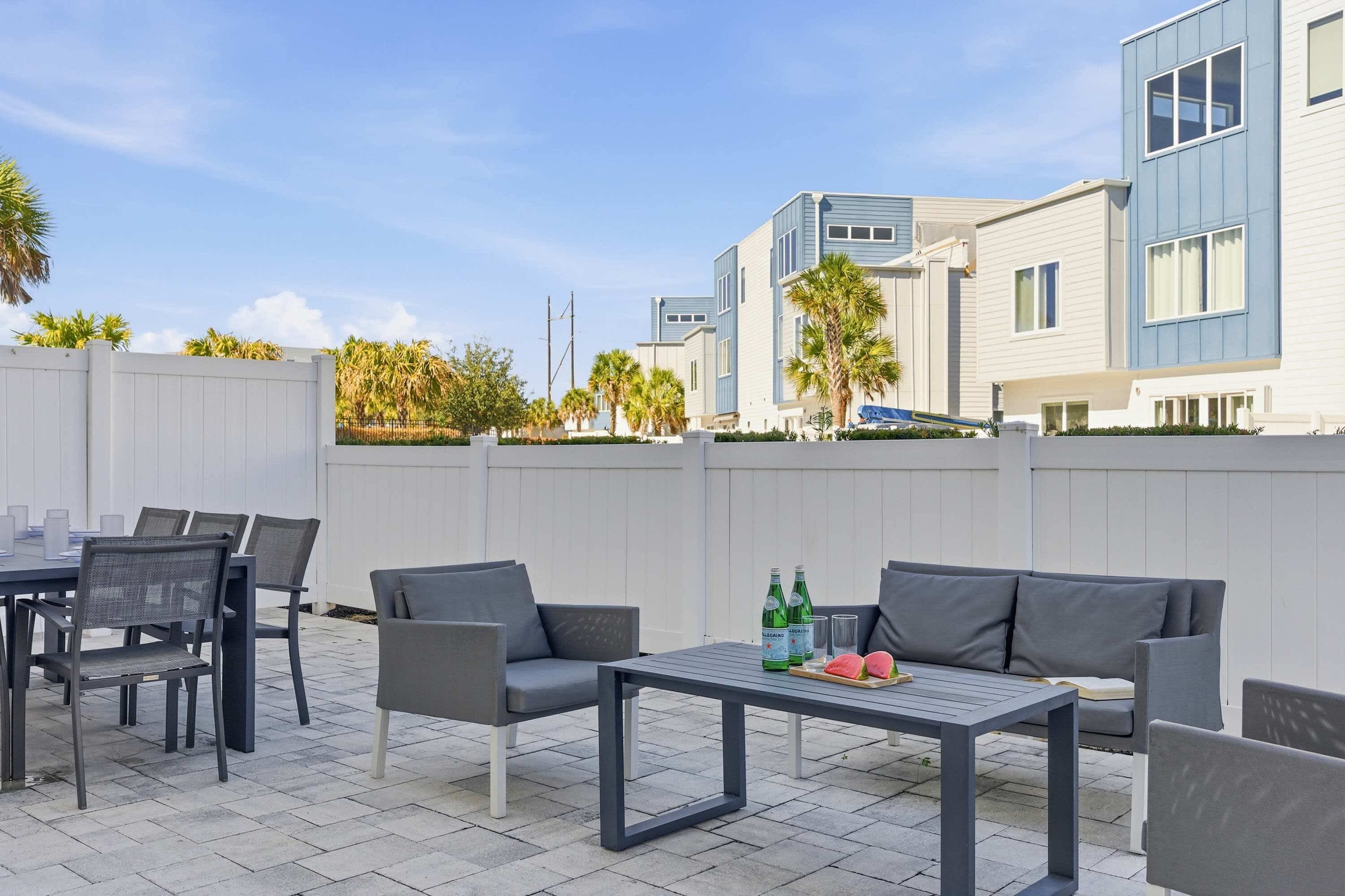 Een terras buiten met een tafel en stoelen, een bank en een bijzettafeltje met flessen en een watermeloen.