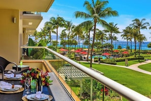 A balcony with a dining setup overlooking a pool area with palm trees and a view of the ocean. - Honua Kai | Ocean View 8 BR, Sleeps 26 | Car Incl. w/ 6+ Nights | HKK ML-3179 by KBM (Lahaina)