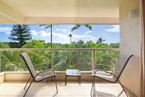 A balcony with two chairs and a small table, overlooking a lush garden with palm trees and a clear sky. - 2BR unit w/ pool & beach access (Kihei)