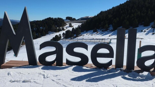 Grandi lettere all'aperto che riportano la scritta 'Masella' su una piattaforma in legno, con un paesaggio innevato e colline boschive sullo sfondo.