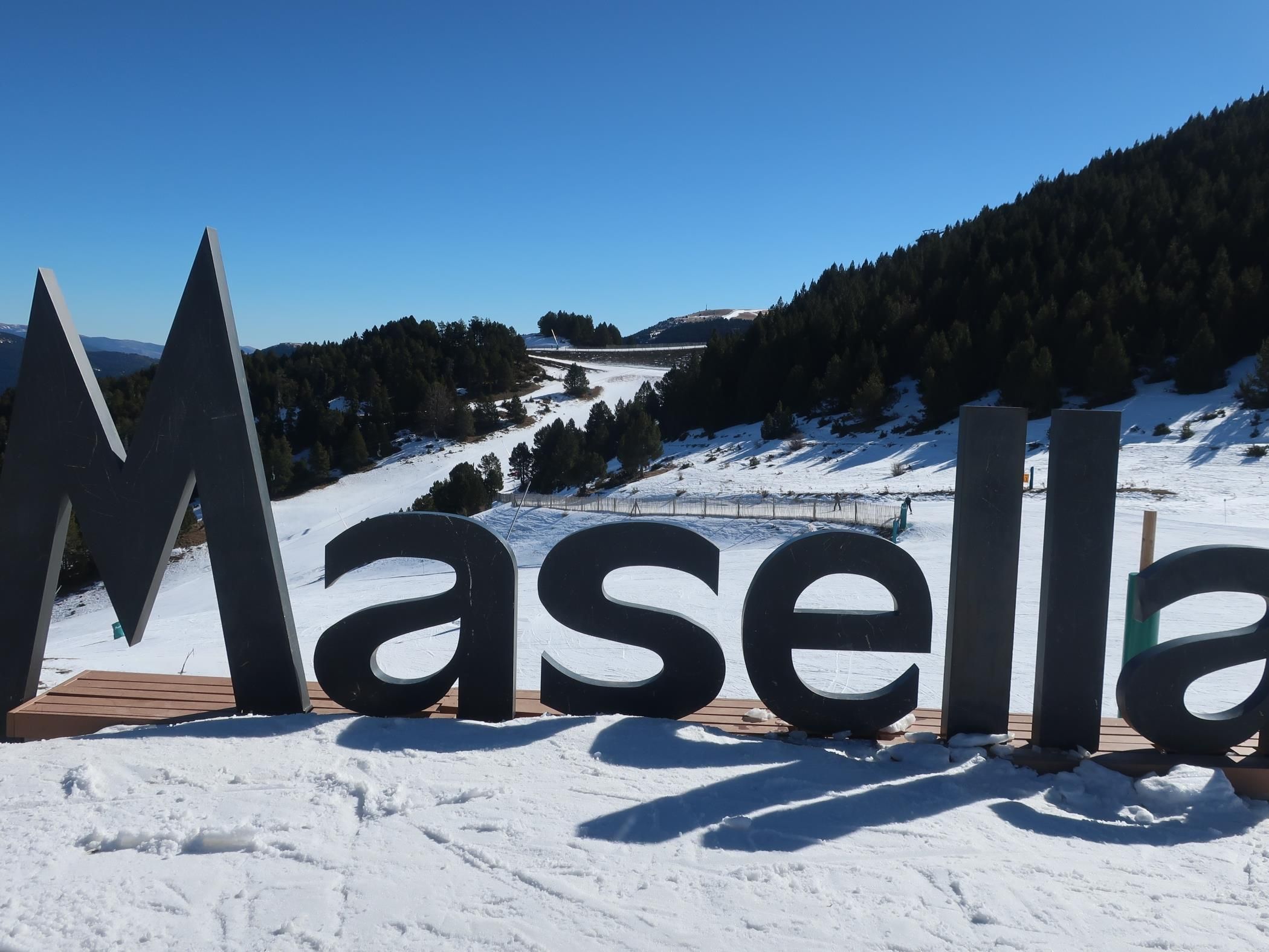 Grandes letras negras que deletrean “Masella” sobre una plataforma de madera con una montaña nevada al fondo.