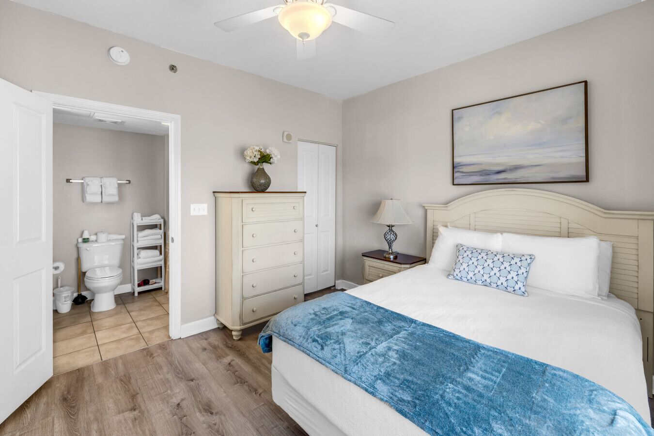 A bedroom with a bed, a nightstand, a dresser, a painting, and a bathroom visible through an open door.