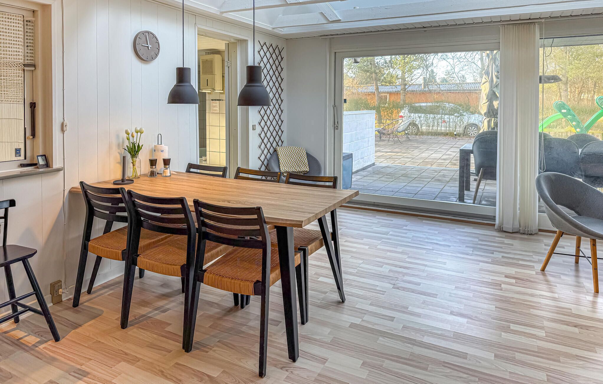 A dining area with a wooden table and chairs, a clock on the wall, and a view of the outdoors through a glass door.