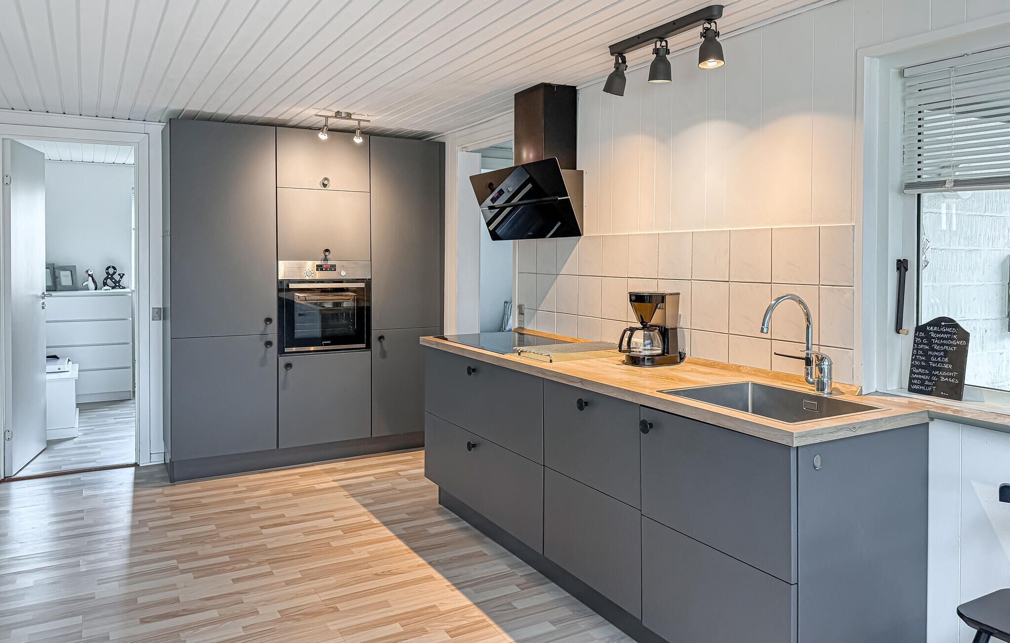 A modern kitchen with grey cabinets, a wooden countertop, a built-in oven, a coffee maker, and a window with blinds.