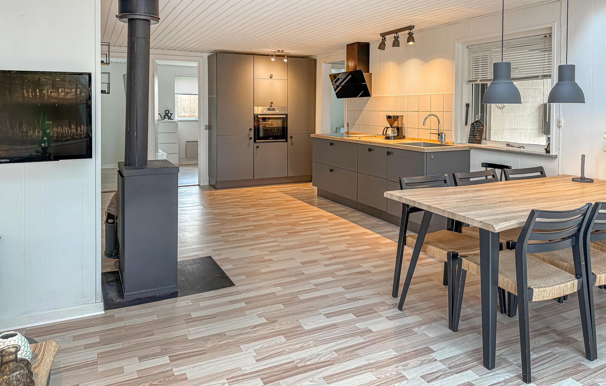 Une cuisine moderne dotée d’un four encastré, d’un coin repas avec une table et des chaises en bois, et d’une grande fenêtre avec des stores.