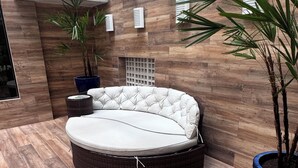 Un espacio de descanso al aire libre con asientos acolchados, base de mimbre, suelo de madera y plantas en macetas.