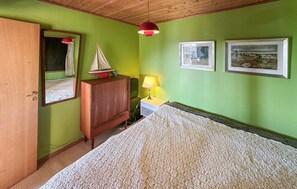 Une chambre avec une armoire en bois, un lit recouvert d’une couverture blanche à motifs, une petite maquette de voilier, une lampe suspendue rouge et deux tableaux encadrés accrochés au mur.
