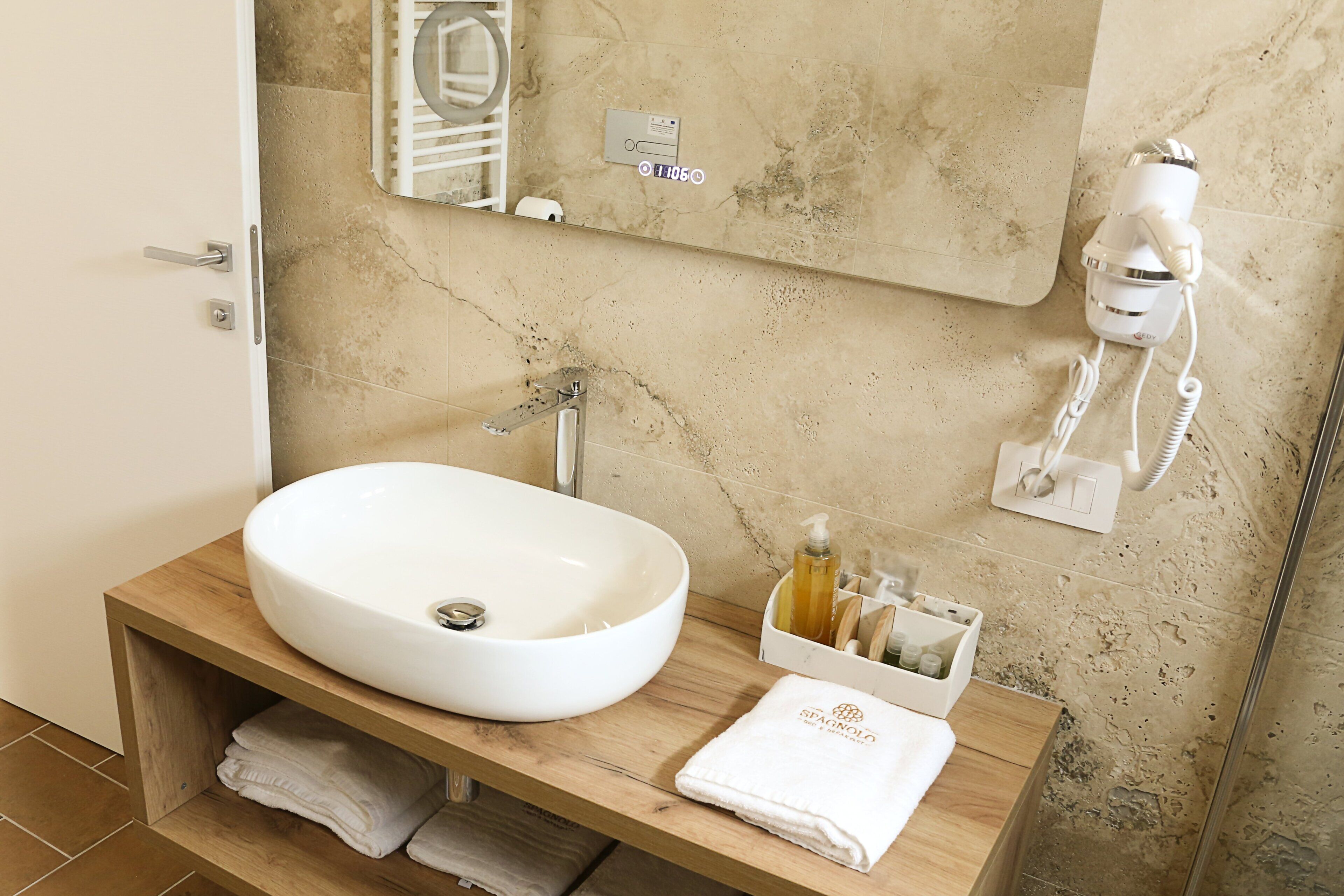 A bathroom with a white sink, wooden vanity, a towel rack, a mirror, a hairdryer, and a soap dispenser.