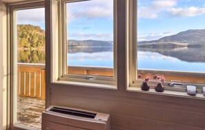Ein Zimmer mit großem Fenster, das auf einen See und Hügel blickt, einer Holzterrasse davor und einem kleinen Heizgerät auf dem Boden.