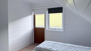 Een kleine, smalle kamer met witte wanden, een houten deur, een raam met een witte omlijsting en een bed met witte lakens.