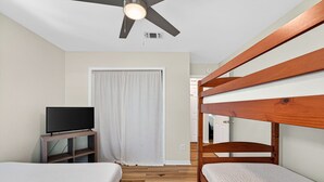 Una habitación con dos camas, un televisor sobre un soporte, un ventilador de techo y una puerta con cortina.