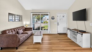 Una sala de estar con un sofá de cuero marrón, una mesa de centro blanca, un televisor sobre un mueble blanco y un ventanal corredizo que da a un balcón.