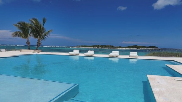 Une piscine d’un bleu éclatant, avec des marches menant à l’intérieur, entourée de chaises longues blanches et de palmiers, offrant une vue sur l’océan et une île lointaine.