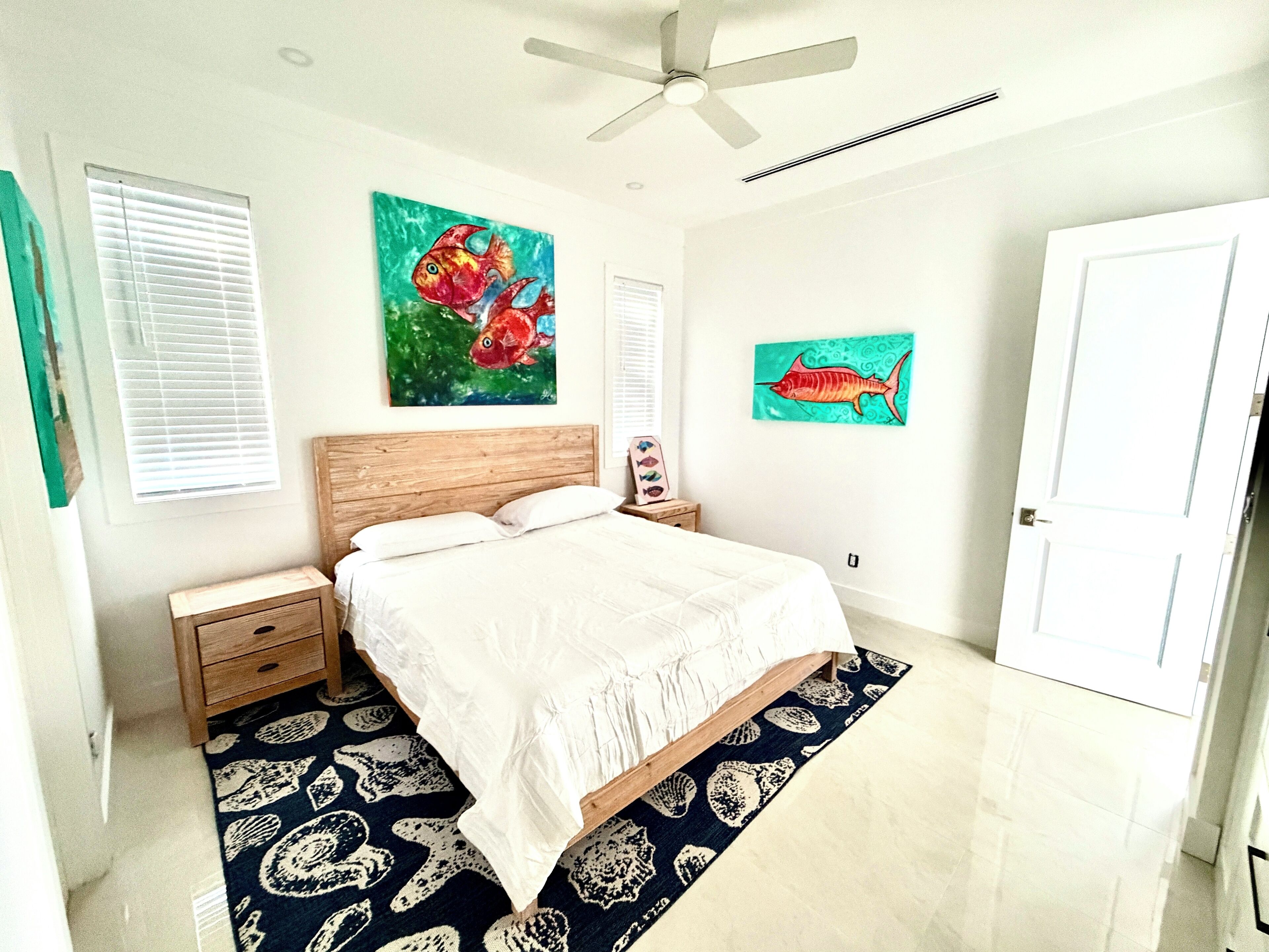 Quarto com cama, ventoinha no teto, duas pinturas penduradas na parede, mesa de cabeceira e uma porta.