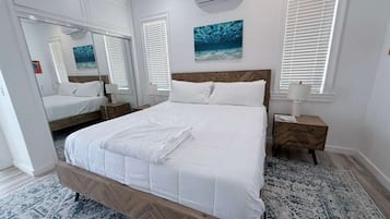 Un dormitorio con estructura de cama de madera, ropa de cama blanca, una mesita de noche, aire acondicionado de pared y un ventanal con persianas.