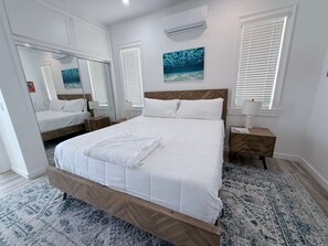 A bedroom with a wooden bed frame, white bedding, a nightstand, a wall-mounted air conditioner, and a large window with blinds.