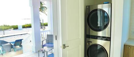 A laundry room with two stacked washers and dryers, a door leading to a balcony with chairs, and a view of the beach.