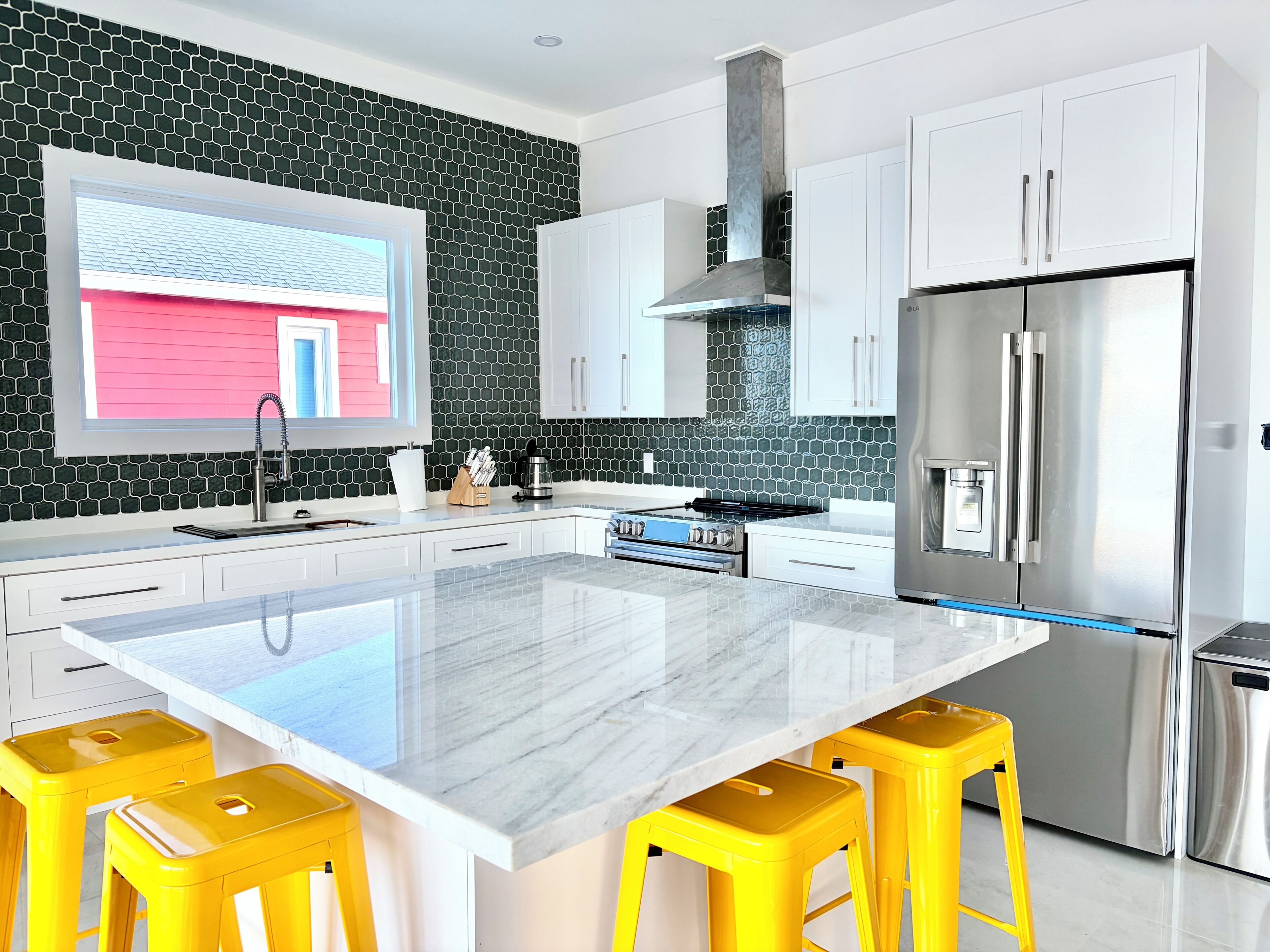 A modern kitchen with white cabinets, a stainless steel refrigerator, a central island with stools, and a window with a view of a pink building.