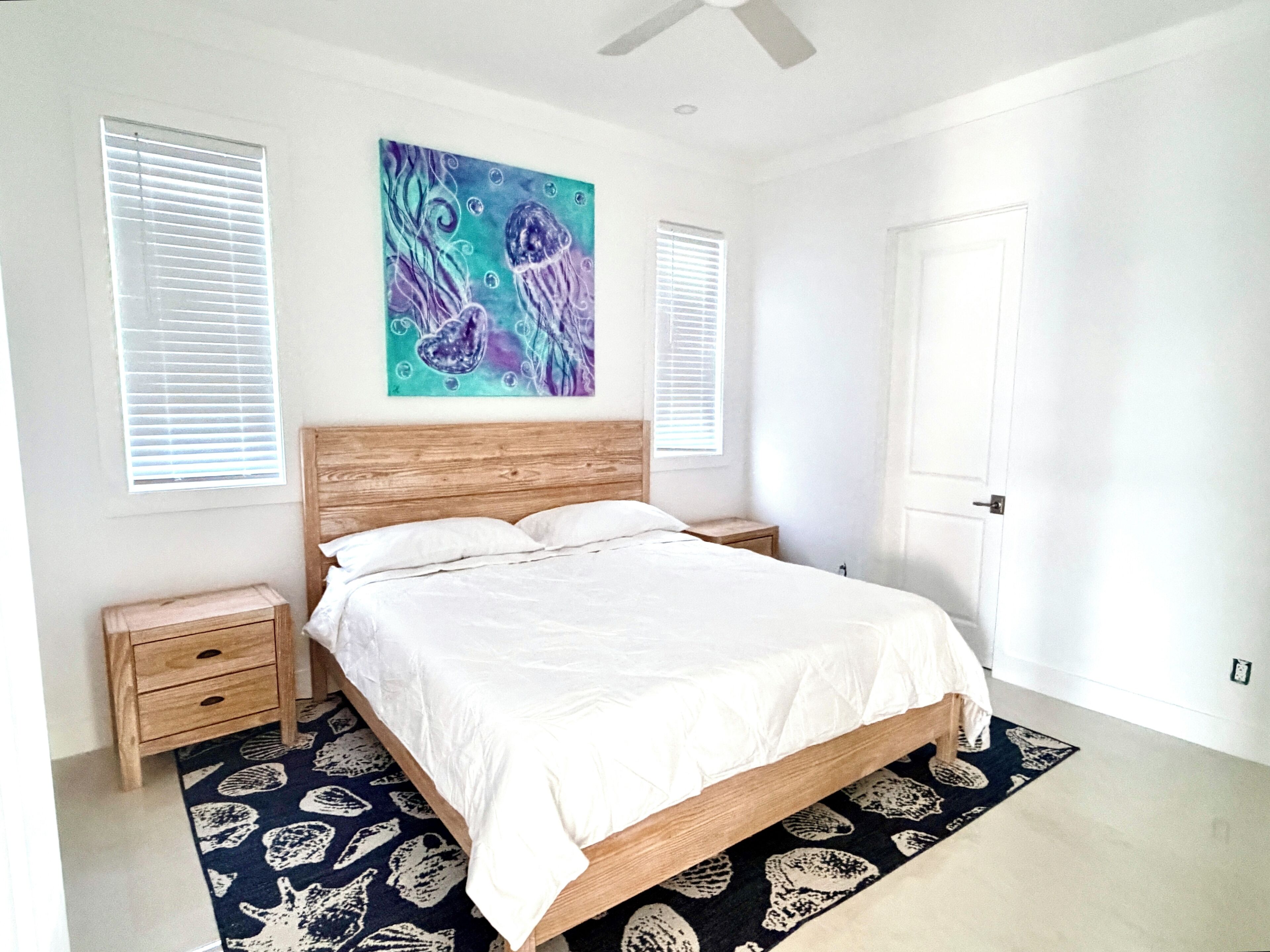 A bedroom with a wooden bed, white bedding, a blue and purple painting, two bedside tables, and a patterned rug.
