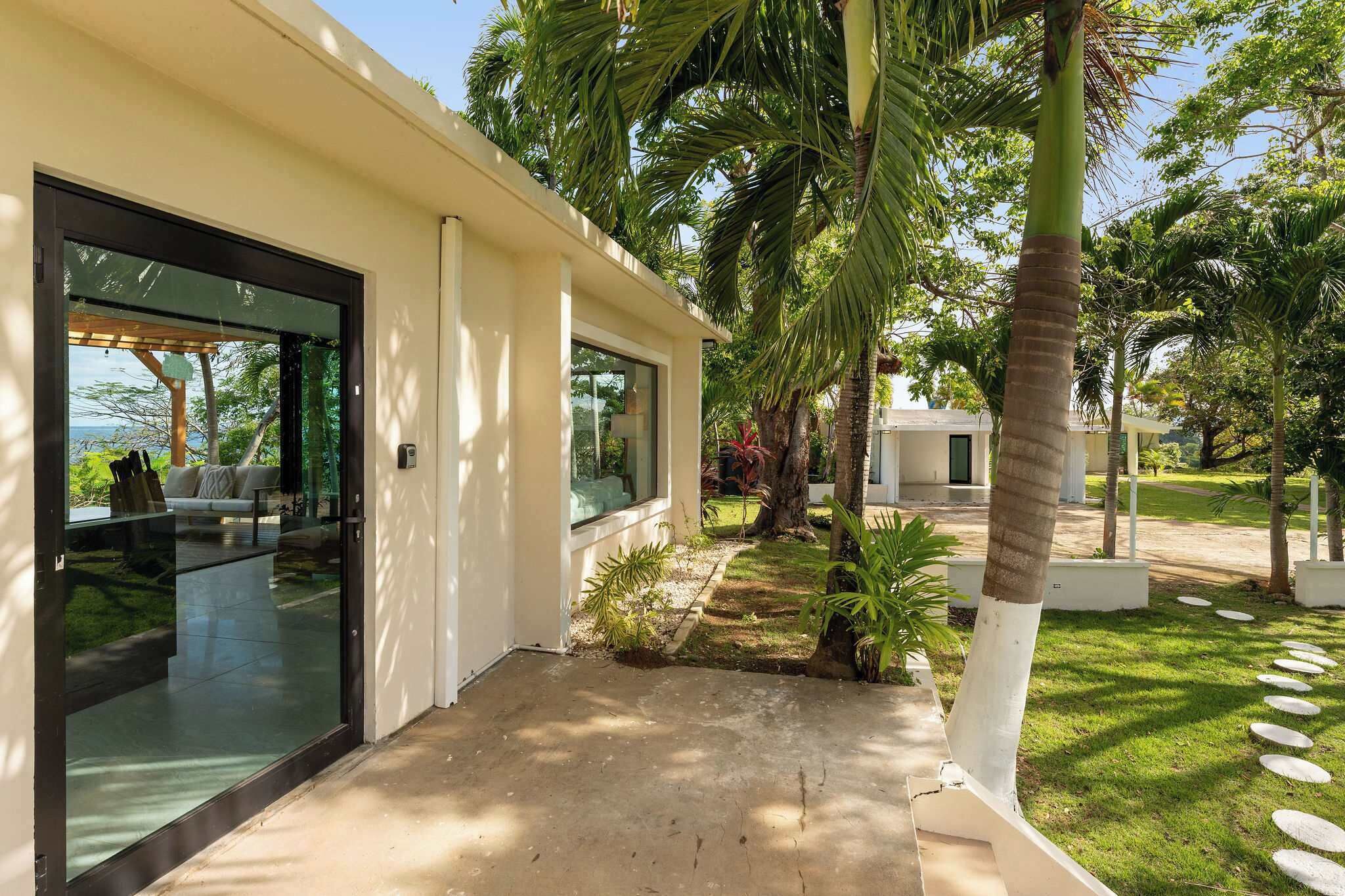 A modern building with large glass doors, a concrete pathway, and a well-maintained garden with palm trees and stepping stones.
