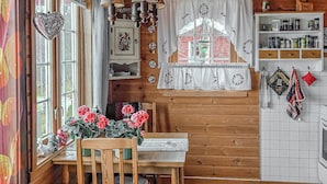 Une cuisine chaleureuse avec des murs en bois, une table en bois, une chaise, une fenêtre avec des rideaux et une étagère remplie d’ustensiles de cuisine.