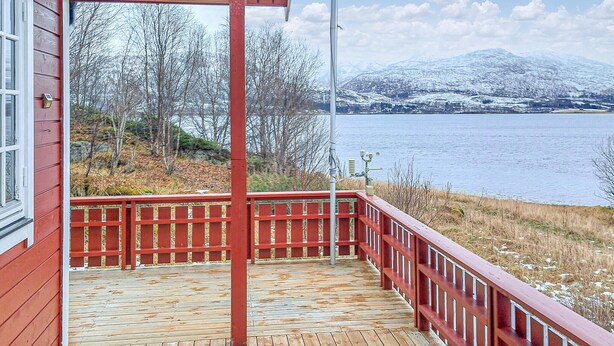 Une terrasse en bois avec une rambarde rouge, donnant sur un lac et offrant une vue sur des montagnes enneigées en arrière-plan.