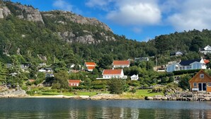 En kustby med hus som ligger nära en vattenyta, omgiven av grönska och en bergig kulle.