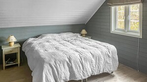 Ett sovrum med ett sluttande tak, en säng med ett vitt överkast, ett sängbord med en lampa och ett fönster med utsikt över träd.