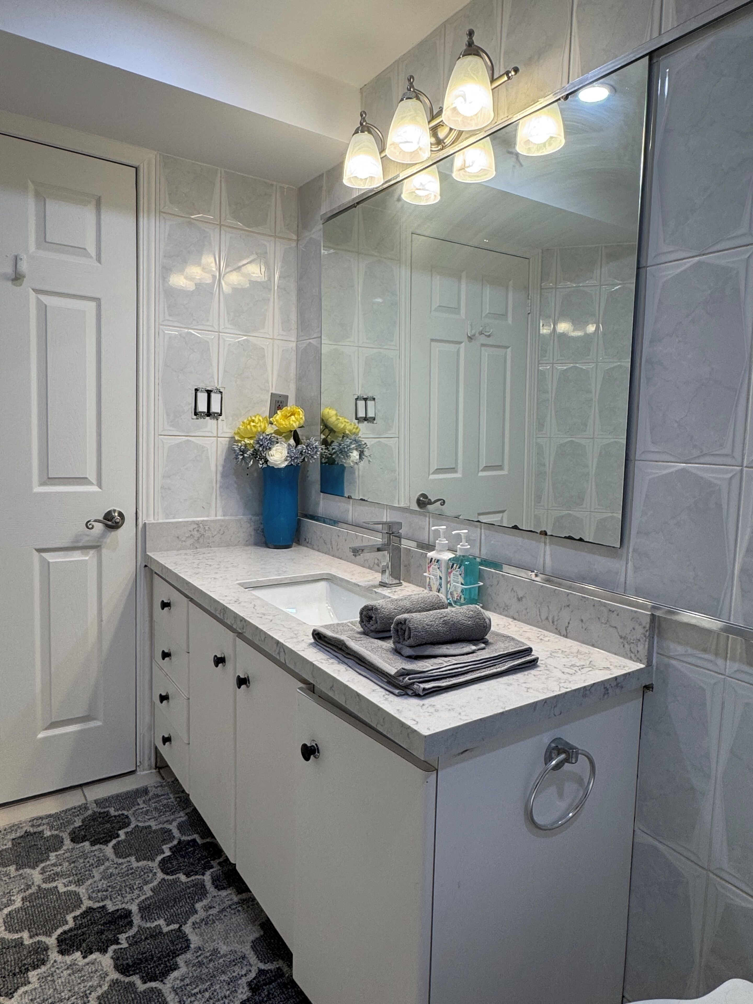 A bathroom with a white door, a marble countertop, a sink, a mirror, and a towel rack.