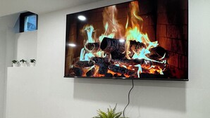 A flat-screen TV mounted on a wall displaying a fire scene, a small table with a plant and remote, and a shelf with decorative items.