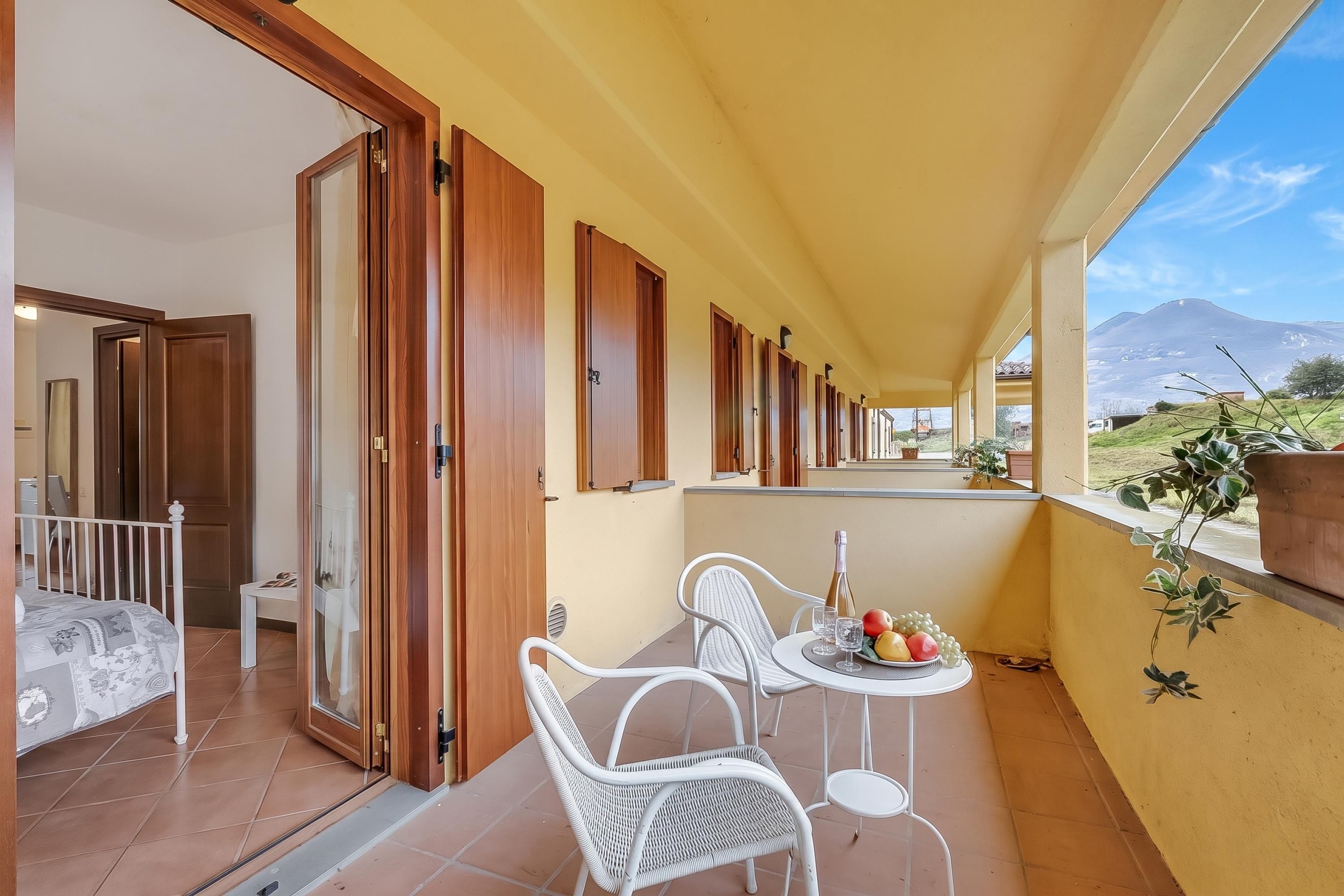 Ein Balkon mit einem Bett, zwei Stühlen, einem Tisch mit einer Flasche und Obst, und Blick auf einen Berg.
