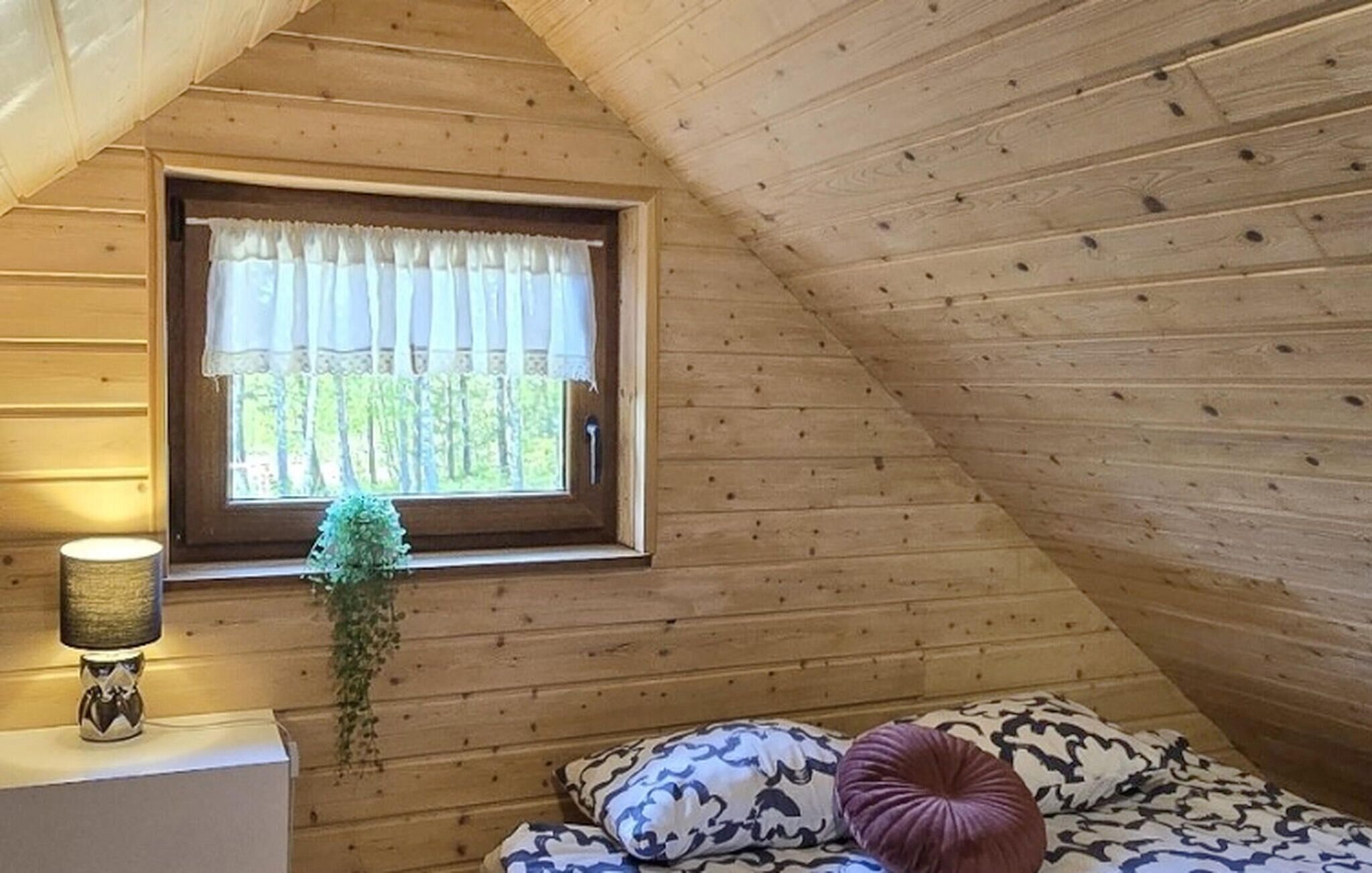 A cozy attic bedroom with a sloped ceiling, a window with a white curtain, a bedside lamp, and a bed with patterned pillows.