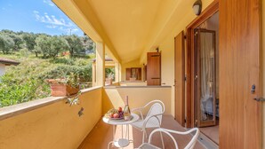 A balcony with two white wicker chairs, a small table with a bottle and fruit, and a view of a garden and buildings in the background. - Residence La Perla - Apt 42, Monte Isola, Italy (Monte Isola)