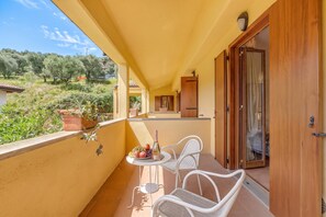 Ein Balkon mit zwei weißen Korbstühlen, einem kleinen Tisch mit einer Flasche und Obst, und Blick auf einen Garten sowie Gebäude im Hintergrund.