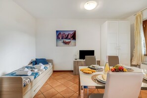 A compact room with a bed, a dining table with fruit and a bottle, a TV, and a painting of a boat on a beach. - Residence La Perla - Apt 33, Monte Isola, Italy (Monte Isola)