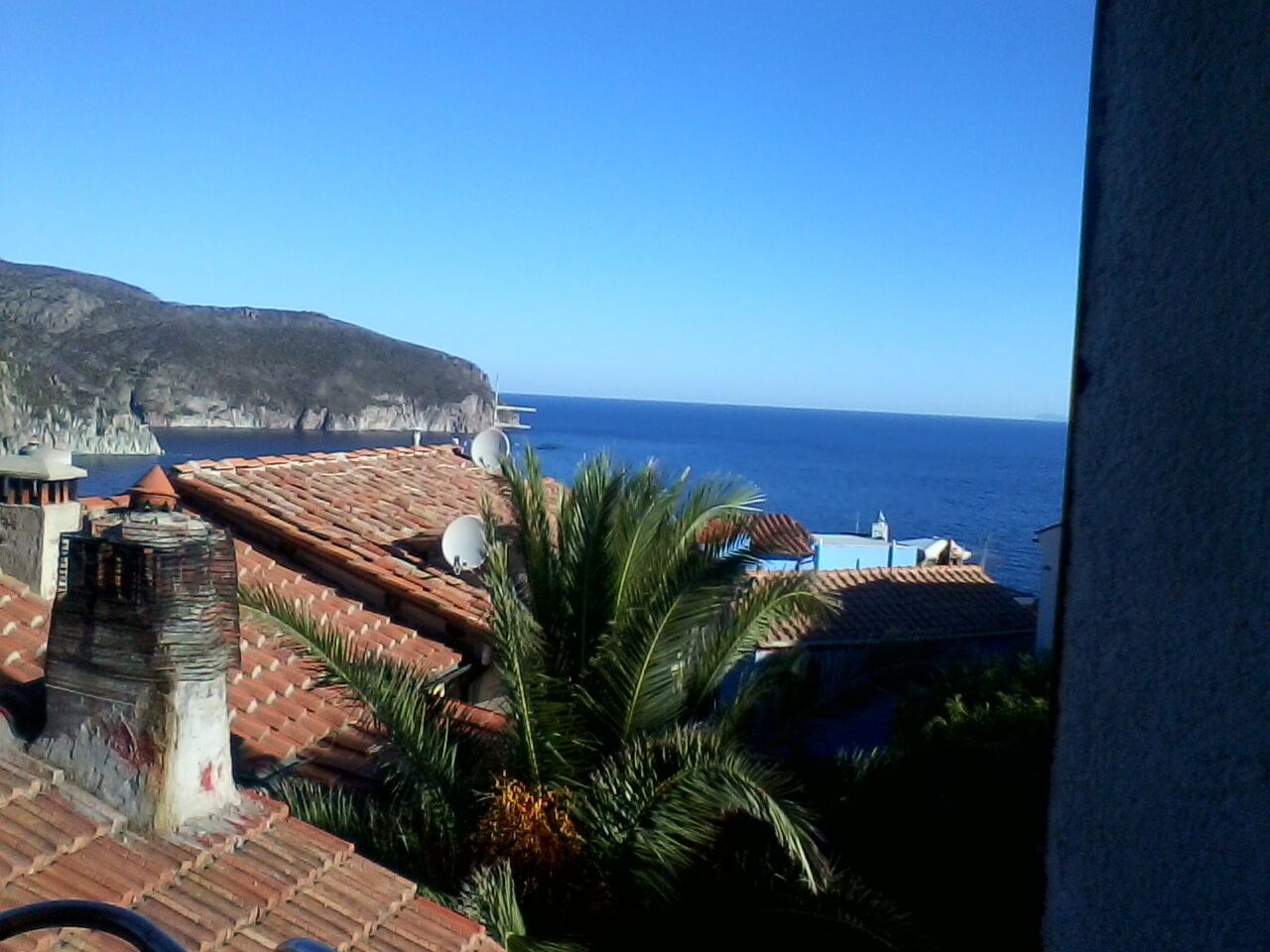 Vista sulla costa con tetti in terracotta, antenne paraboliche e cielo azzurro limpido.