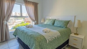 Un dormitorio con cama, dos almohadas, una mesita de noche con lámpara y una ventana con cortinas.
