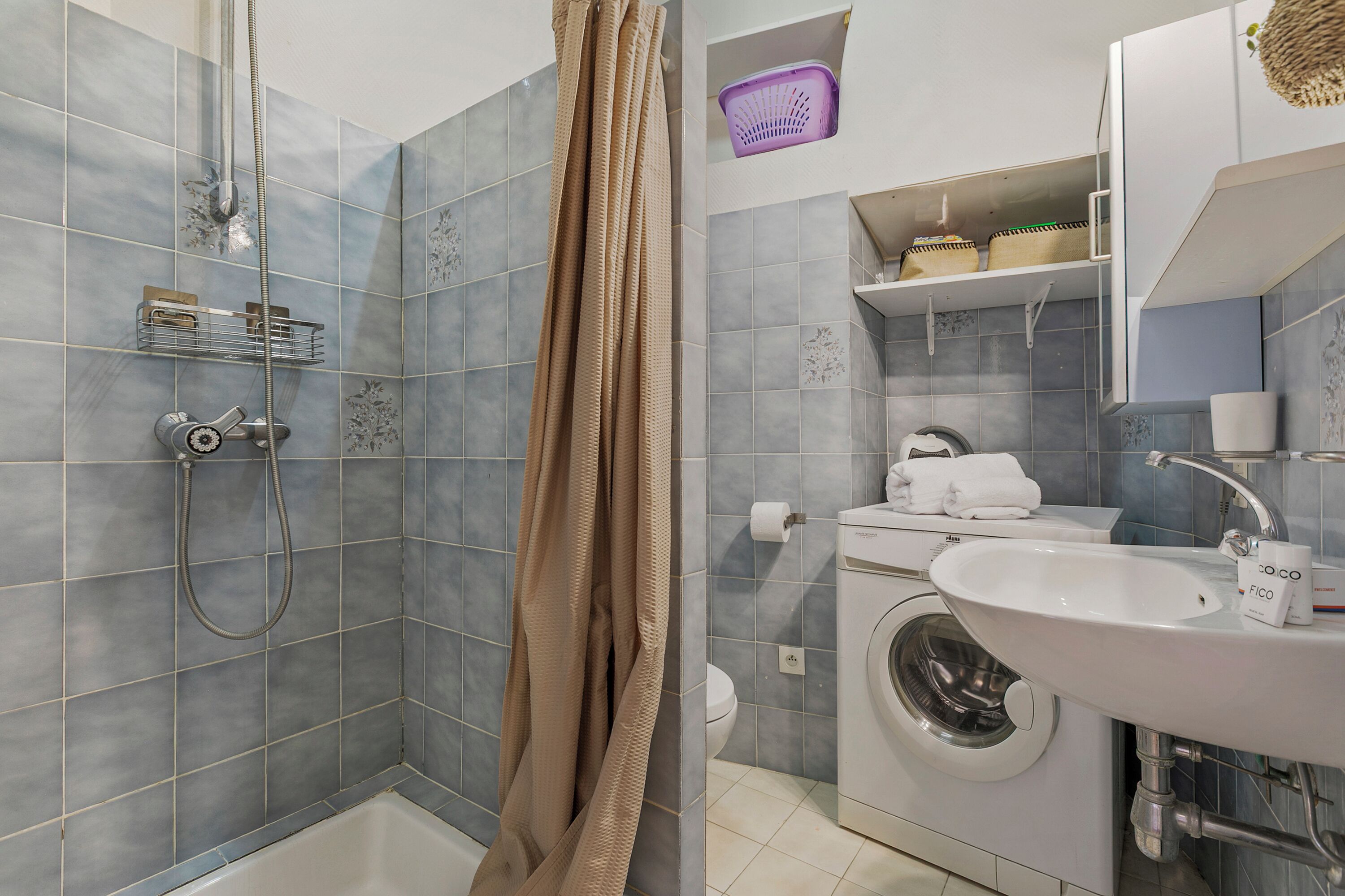 A bathroom with a shower, toilet, washing machine, sink, and storage shelves.