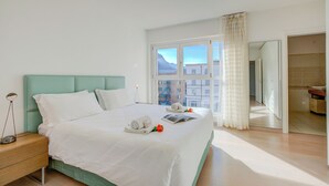 Une chambre d’hôtel moderne avec un grand lit, une table de chevet avec une lampe, une fenêtre donnant sur des immeubles et une salle de bain visible à travers une porte ouverte.