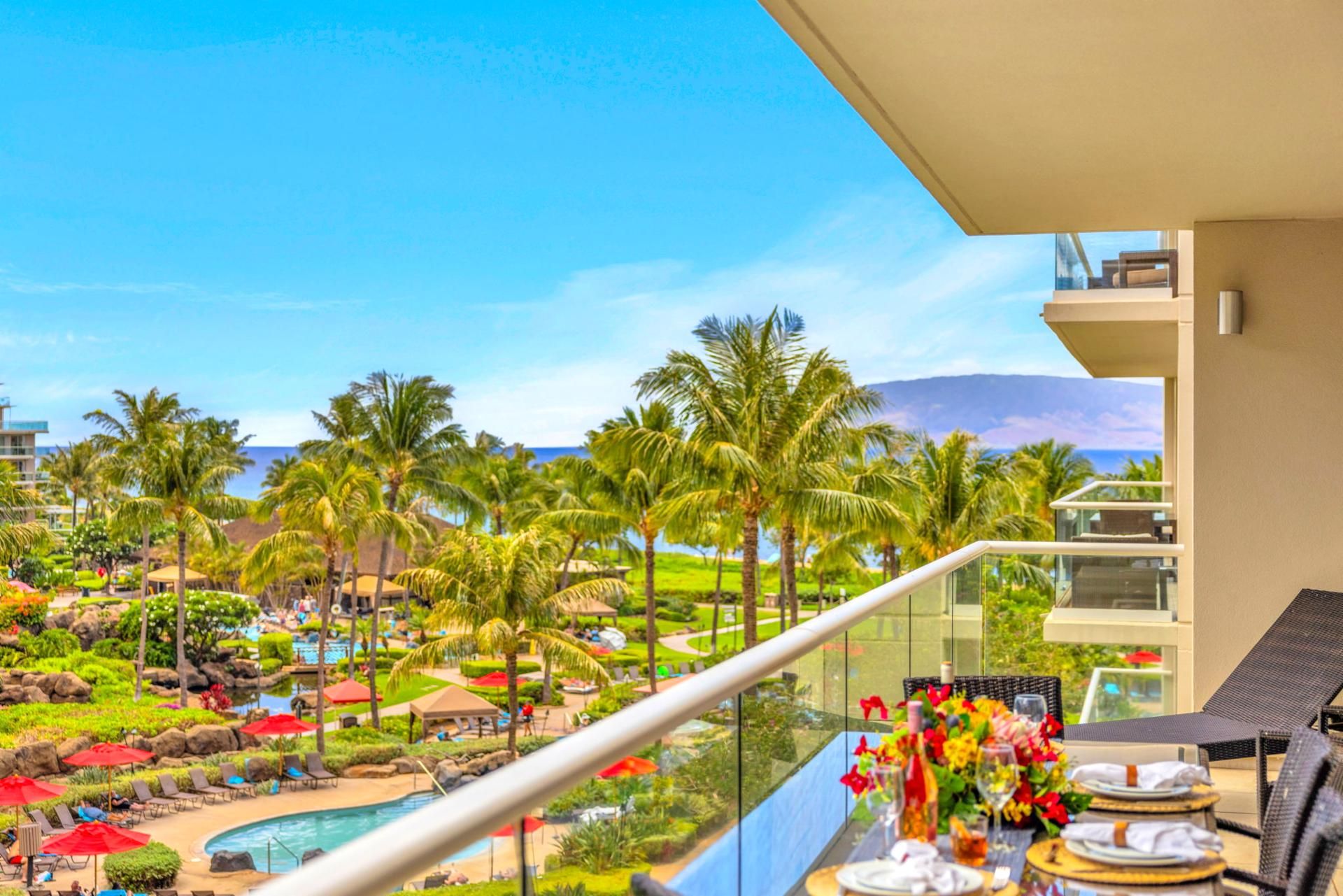 Un balcón con barandilla de cristal que da a una piscina del complejo turístico, palmeras y un paisaje montañoso.