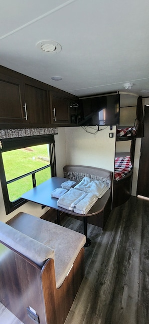 A compact RV interior with a kitchenette, a small dining table, a sleeping area, and a television.