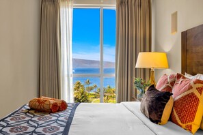Ein Hotelzimmer mit großem Fenster, einem Bett mit gemusterten Betttextilien, einer Lampe und Blick aufs Meer.