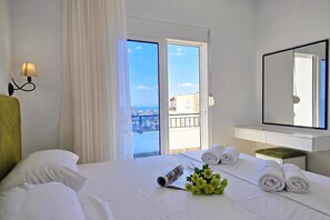 A hotel room with a bed, a bedside table with towels, a window with a view of buildings, and a wall-mounted lamp. - Ionian Blue Escape 304 /w Sea View Balcony, Sarandë, Albania (Sarandë)