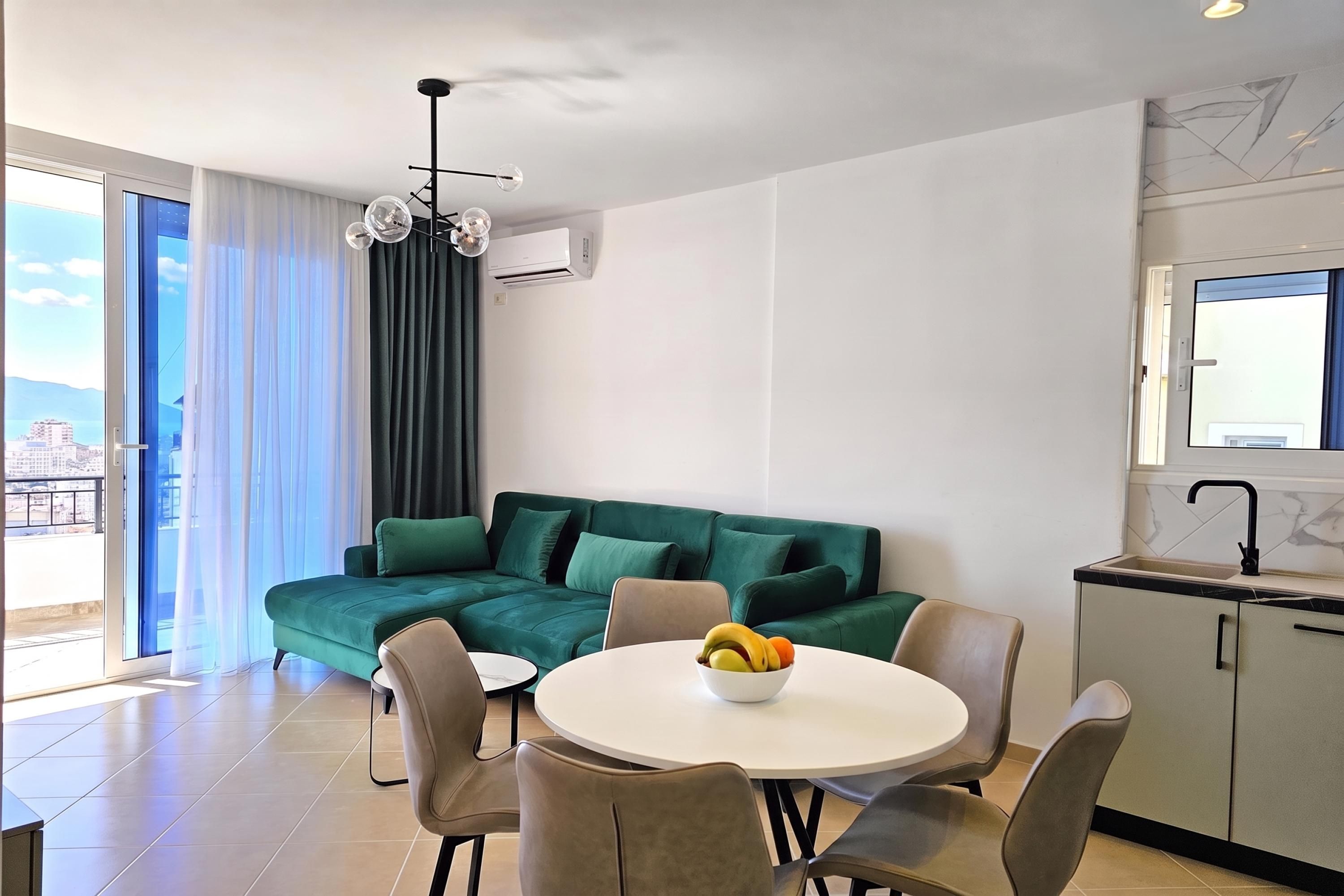 A modern living room with a green sofa, a dining table with chairs, and a kitchen area with a sink and window.
