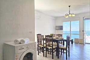 Ein Zimmer mit Waschmaschine, einem Esstisch mit Stühlen, einem Fernseher und Meerblick durch große Fenster.