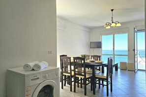 A room with a washing machine, a dining table with chairs, a chandelier, and a view of the sea through large windows. - Two Bedroom Apartment With Sea View-061, Sarandë, Albania (Sarandë)