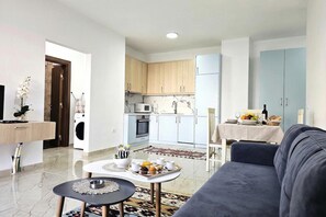 A modern living room with a sofa, a coffee table with a tray of food, a TV, and a kitchen in the background. - One Bedroom Apartment In Saranda - 001, Sarandë, Albania (Sarandë)