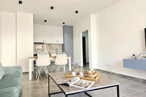 A modern living room with a sofa, a coffee table with a magazine, and a kitchen in the background. - Holiday Apartment In Saranda - 003 With Balcony, Sarandë, Albania (Sarandë)