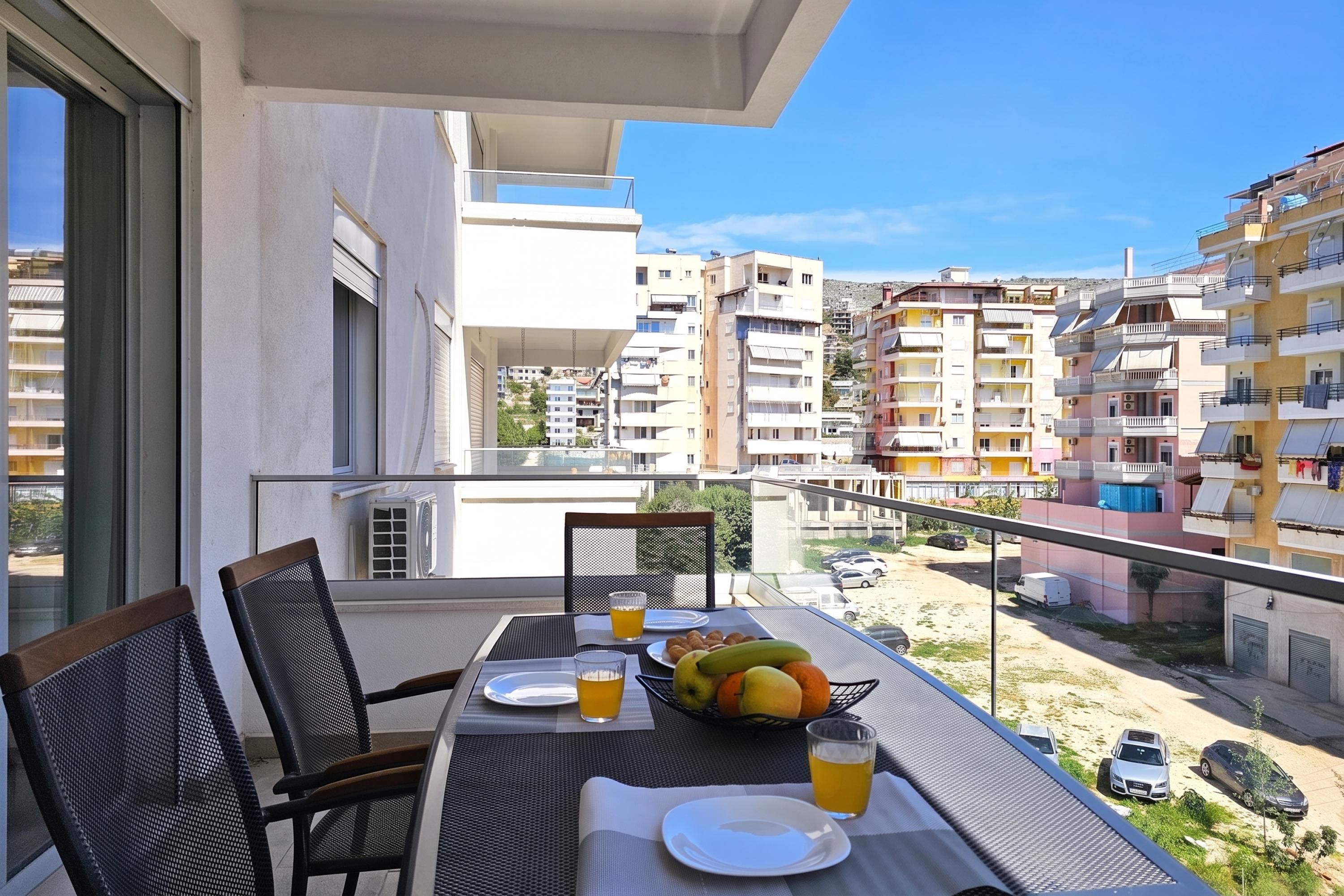 A balcony with a table set for two, chairs, a fruit bowl, and a glass of orange juice.