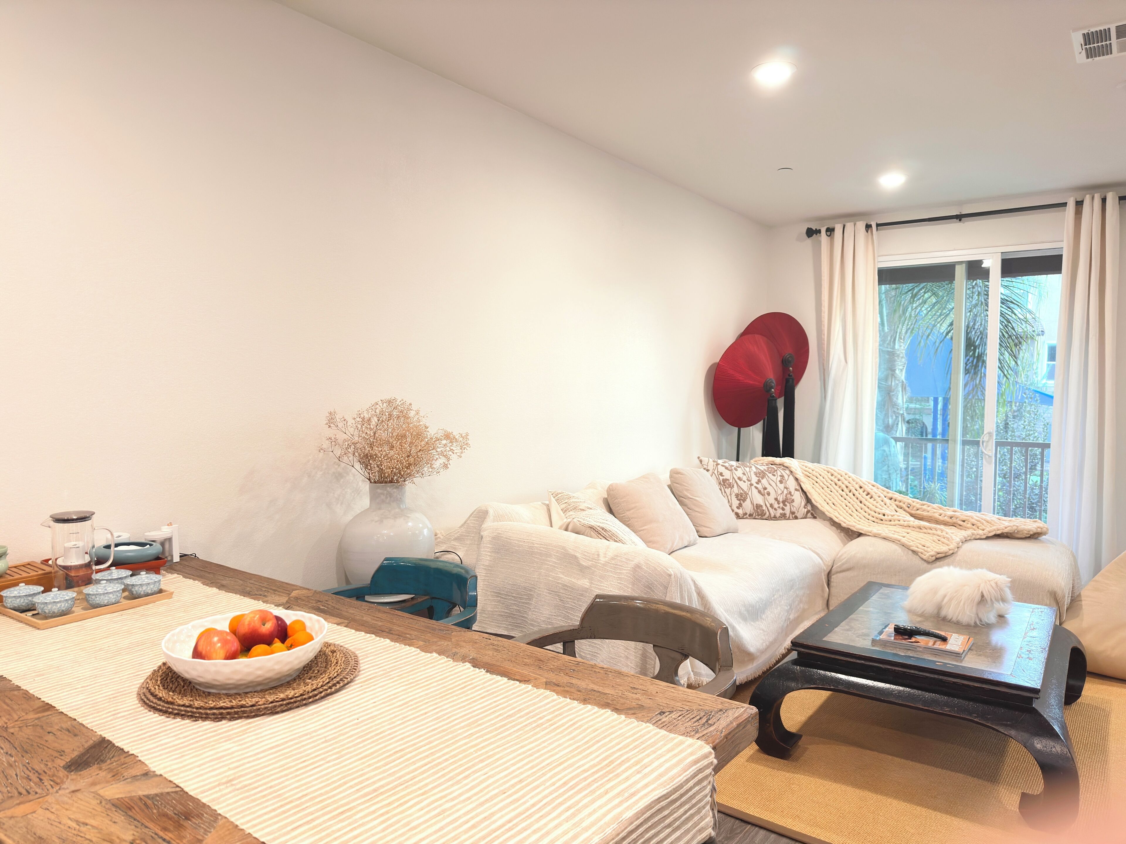 A living room with a sofa, a coffee table, a dining table with a bowl of fruit, a vase with dried flowers, and a window with curtains.